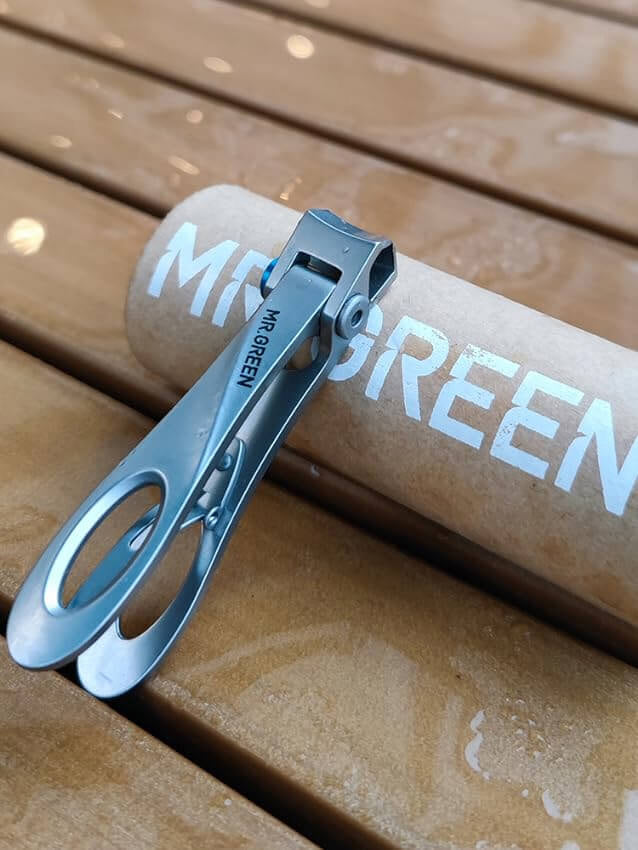 MR.GREEN wide-jaw stainless steel toenail clippers for thick nails with ergonomic loop handle, lifestyle photo on wet wood beside branded tube